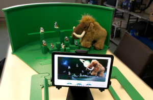 Greenscreenbox mit in Szene gesetzten Ü-Eierfiguren und Stoffmammut. Im Vordergrund sieht man ein tablet mit Weltallszenerie und den Figürchen.